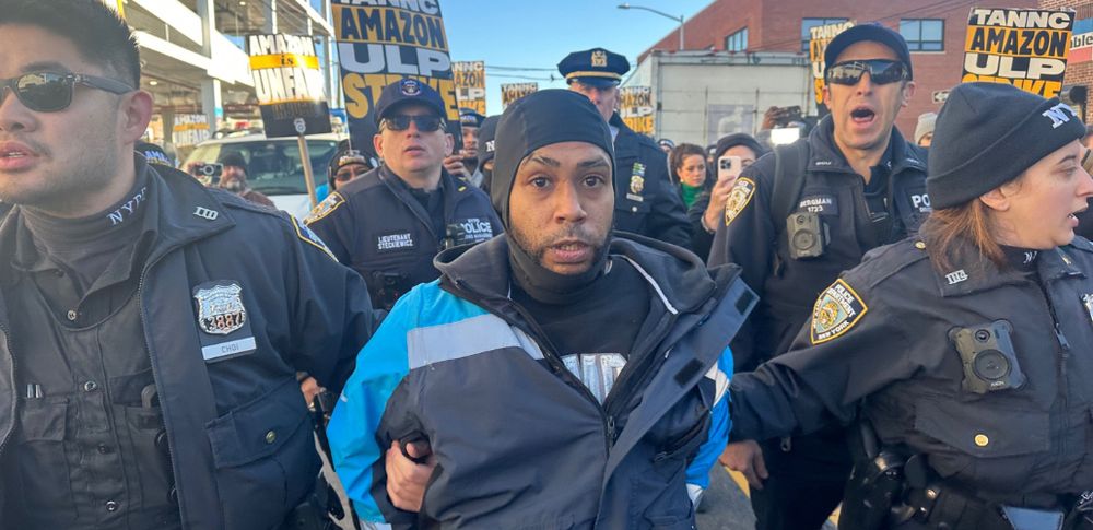 Jogernsyn Cardenas, an Amazon delivery driver, is arrested by NYPD at an Amazon strike. He is cuffed and escorted by 2 police officers, with 3 officers behind him. 
