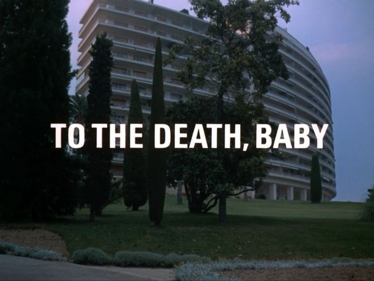 Titlecard from the television program THE PERSUADERS. In colour, what appears to be a Postwar Modern International-styled apartment building seen from a short  distance at a slight lower angle across an expanse of close-cropped green grass.

Centred in the frame's top two thirds, it's a gently-curving white concrete structure supported by innumerable narrow pilotis at a ground level concourse. The building's left terminal end has ten storeys; as it horizontally arcs away to the right of frame, two additional storeys appear at roughly centre frame for a total of twelve floors. Each floor appears to feature a recessed loggia, or terrace, whose continuously curving edges and railings are in fact what constitute the building's face.

Its leftmost side is partially obscured by a clutch of various decorative landscaped trees and shrubs* which, several metres down a slightly sloped grassy lawn, stretch horizontally to the frame's midpoint from its dark left edge. The largest of these, an unidentifiable slim hardwood tree, bisects our view of the building as it rises up and out of frame. 

Under a darkening blue sky, the image's overall dimness suggests dusk. (Absent additional context, it may have been shot day-for-night.) 

Overlaid in a font of weighty white sans serif capitals** on a single vertically-centred line, "To the Death, Baby".


(Reasonably certain this is the Residences Saint Michel-Valetta, 69 rue de roi albert, in Cannes, designed in 1958 by Louis Lafond. It also plays a significant role in Truffaut's Woolrich adaptation THE BRIDE WORE BLACK. Good movie.  

Two additional views -- how it appeared on a picture postcard (undated, though predating 1994), and a more recent photo from a real estate agent -- are available here:

https://imgur.com/a/6SYyS1L

)

*(I'm by no means a horticulturist, but am I seeing a coupla conical boxwoods, there? Maybe a privet tree?)

**(a Helvetica variant (neue?)? Bold condensed Univers 67? Could be either. Or neither!)
