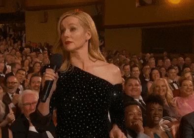 Laura Linney, a white woman with shoulder length blonde hair, red lipstick, and award show makeup, is standing in a sequined off the shoulder black gown in front of a crowd of people with a microphone held out and a look of “what are you gonna do about it?” (If he image moves, she’s making a shrugging motion while the crowd behind her laughs and applauds)