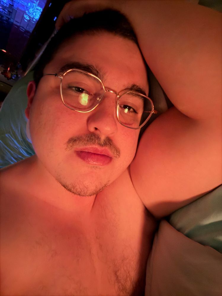 Selfie of me lying shirtless in bed, bathed in a warm light, giving a sly smirk to the cam. My right arm (shown as left in flipped photo) is placed seductively behind my head to further sweeten the deal. 