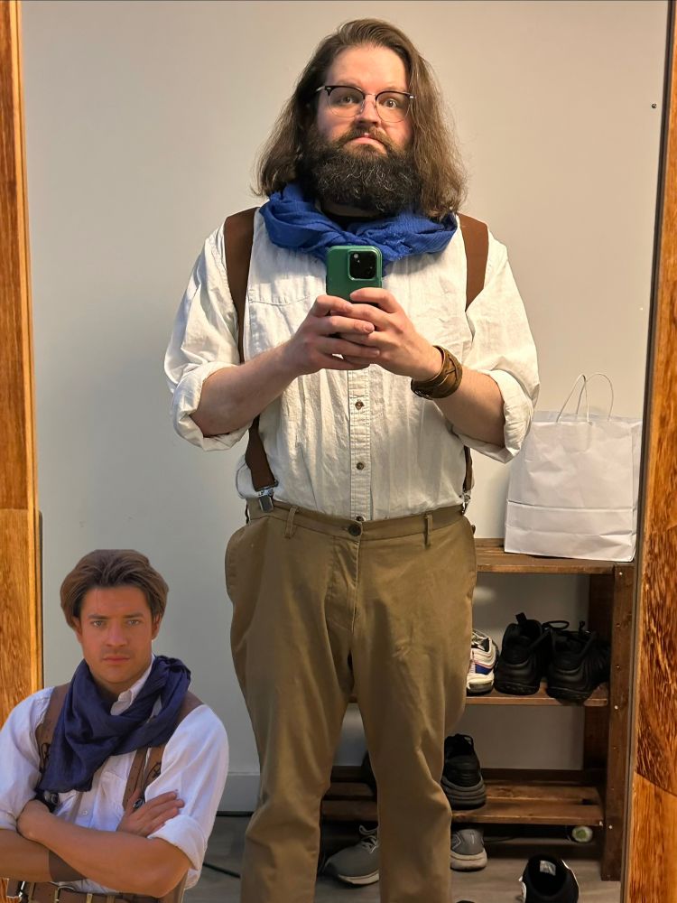 A mirror selfie of a man in white shirt, brown pants, suspenders and a blue scarf. Rick O’Connell from The Mummy is in the bottom corner, arms folded.