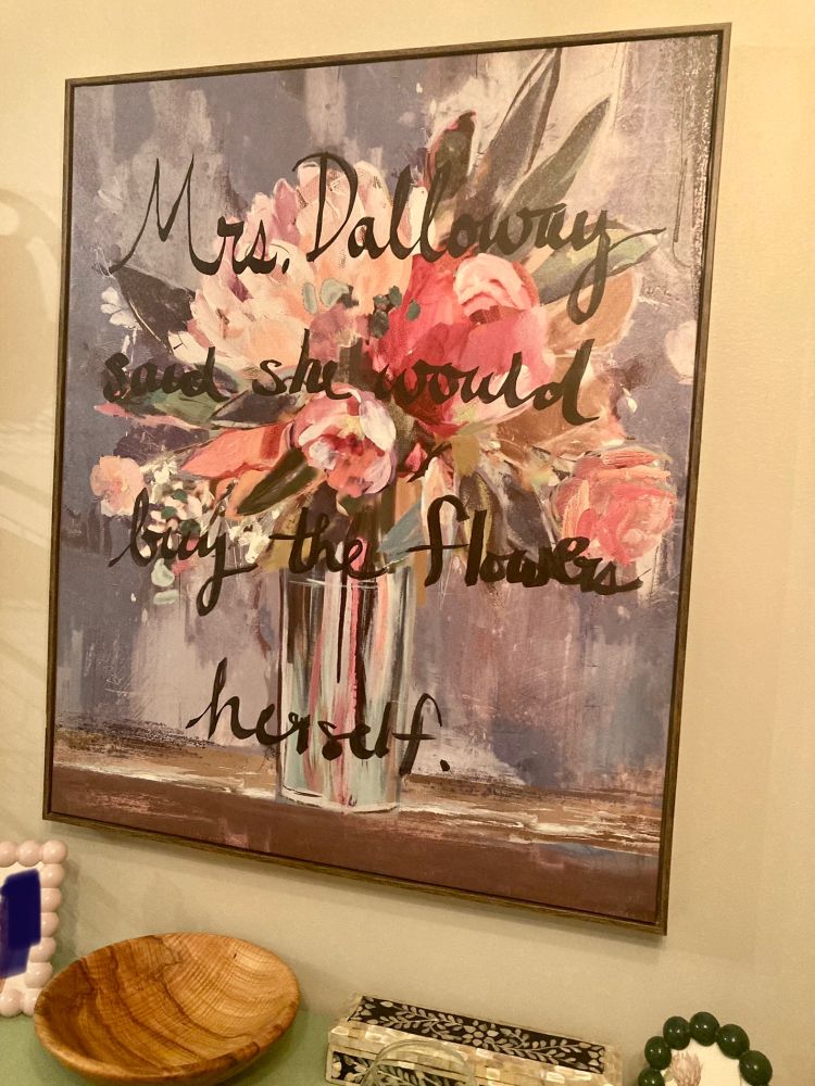 A large painting of flowers in a vase with “Mrs. Dalloway said she would buy the flowers herself” painted on top in black cursive script