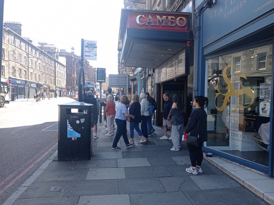 Street Scene with bus stop, passengers and customers going into and out of the Cameo Cinema in Edinburgh.  It has an open porch over the street. 