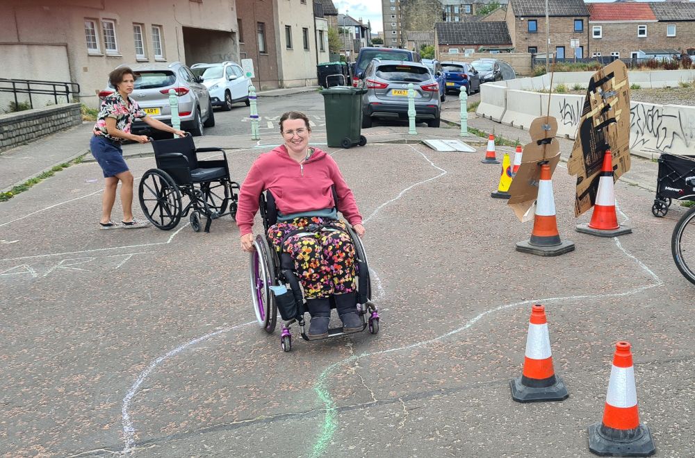 Wheelchair obstacle course, with curvy chalk lines, road cones, and a ramp in the distance.  Person sitting in a wheelchair and person pushing a wheelchair. 