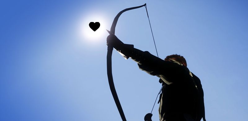 An archer takes aim backlit by the sun. He aims at a black illustrated heart.