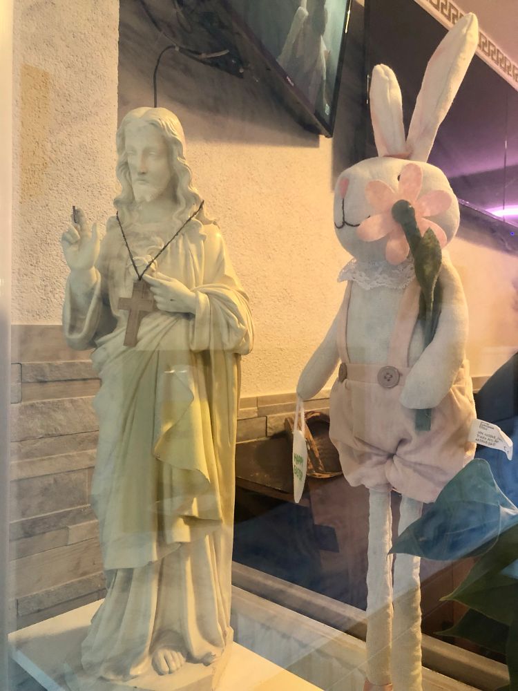 Eine etwa 70 cm hohe, segnende Jesus-Statuette steht in einem Schaufenster. Direkt daneben eine kitschige moderne Osterhasenfigur aus Stoff. 