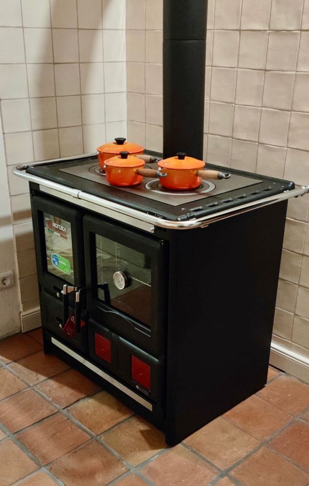 Ein schwarzer Holzküchenofen aus Metall auf Cottofliesen. Es gibt ein großes Backfach rechts einen Brennraum links und oben auf dem Ofen zwei Herdplatten. Das Rauchrohr befindet sich mittig und steigt senkrecht nach oben auf.