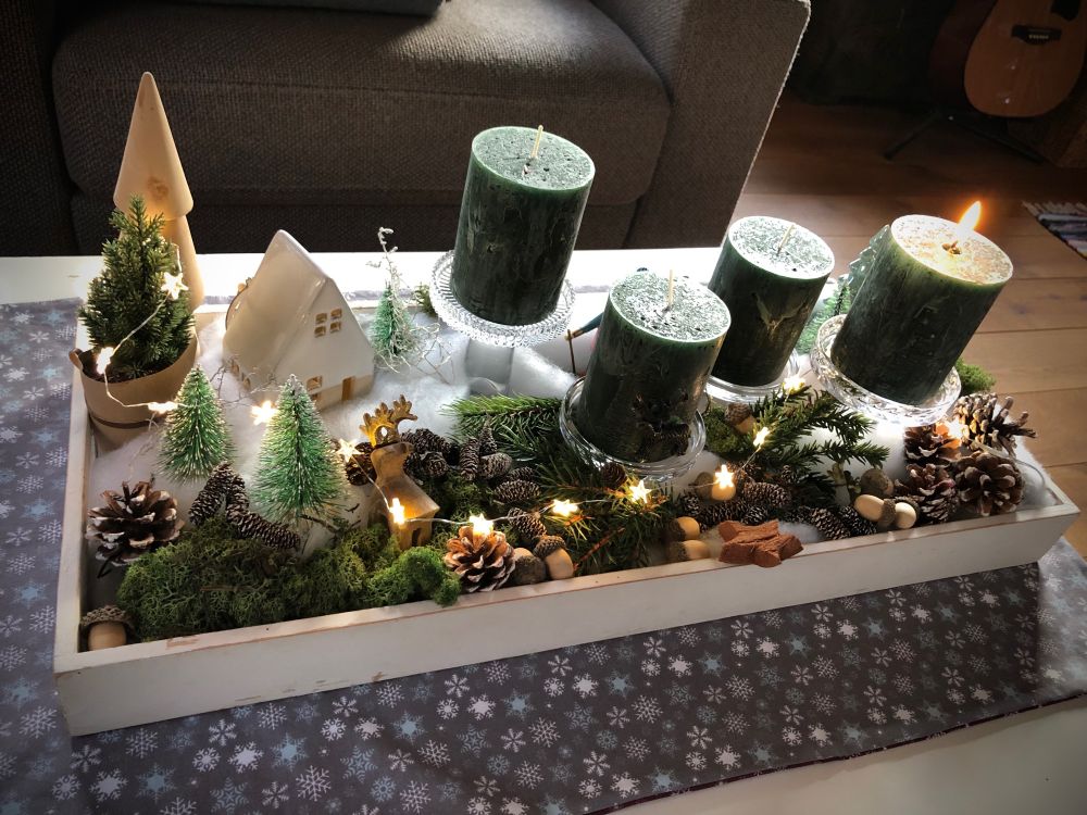 Ein weißes Holztablett mit einer Winterschneelandschaft. Diverse Tannenbäume aus Holz, Porzellan, Kunststoff, ein beleuchtetes Häuschen aus Porzellan, ein Holzhirsch, eine Langläuferin aus Holz. Diverse Zapfen und Eicheln, etwas Moos, eine Lichterkette mit leuchtenden Sternen und vier Kerzenständer aus Glas mit dunkelgrünen Kerzen. Eine Kerze brennt.