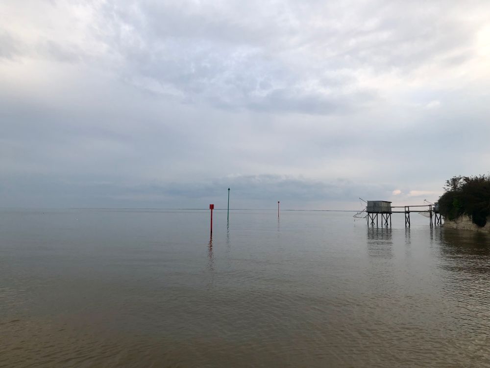 Blick auf ruhiges Wasser. Der Abendhimmel verschmilzt mit dem Horizont. Links eine Fischerhütte auf Stelzen, mittig drei Seezeichen.