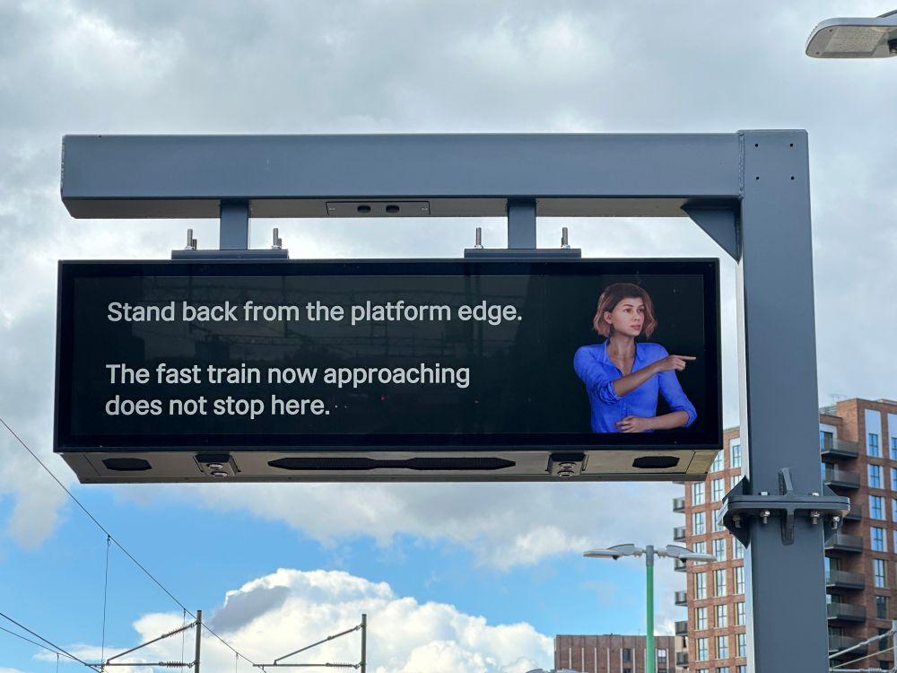 A railway platform indicator board displays the following message: “Stand back from the platform edge. The fast train now approaching does not stop here.” Next to the text is a computer-animated woman relating the message in British Sign Language.