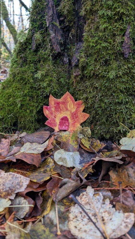 A mossy green tree has a red/orange clay leaf with a tiny red door leaned against it.