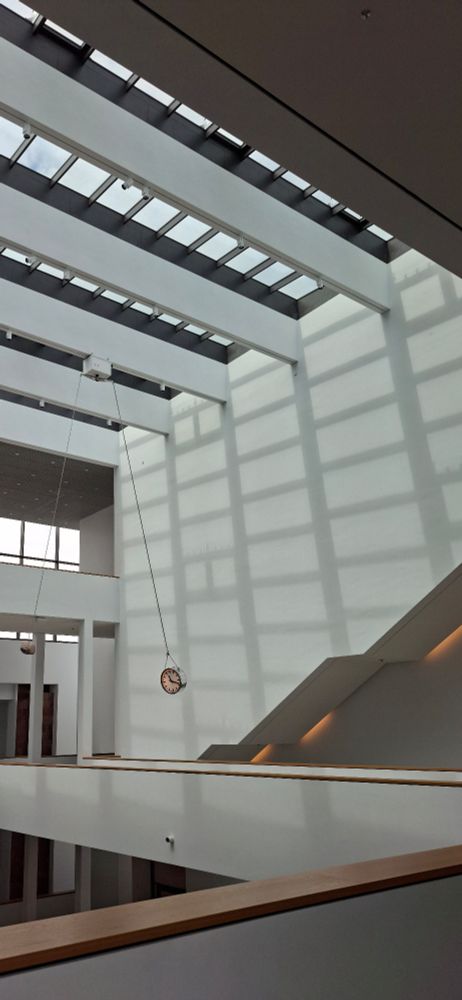 Das Bild zeigt das Innere eines modernen, lichtdurchfluteten Gebäudes mit minimalistischer Architektur. Der Raum ist groß und offen gestaltet, mit weißen Wänden und klaren Linien. Die Decke besteht aus mehreren Glasdachfenstern, durch die Tageslicht einfällt und markante Schatten von den Trägern und Fensterrahmen auf die Wände wirft. An der rechten Seite verläuft eine breite Treppe nach oben. Im Mittelpunkt des Bildes hängt eine große, runde Uhr an einer langen Kette von der Decke herab, an einer zweiten Kette hängt ein Stein mit der ungefähren Größeder Uhr. Die Wände sind glatt und weiß, was den Raum noch heller und offener wirken lässt. An den Brüstungen der oberen Etage verläuft ein Holzhandlauf, der einen warmen Kontrast zur sonst kühlen Farbgebung bietet. Die Atmosphäre wirkt ruhig, geordnet und einladend es handelt  sich um das Foyer der Kunsthalle Mannheim. 

