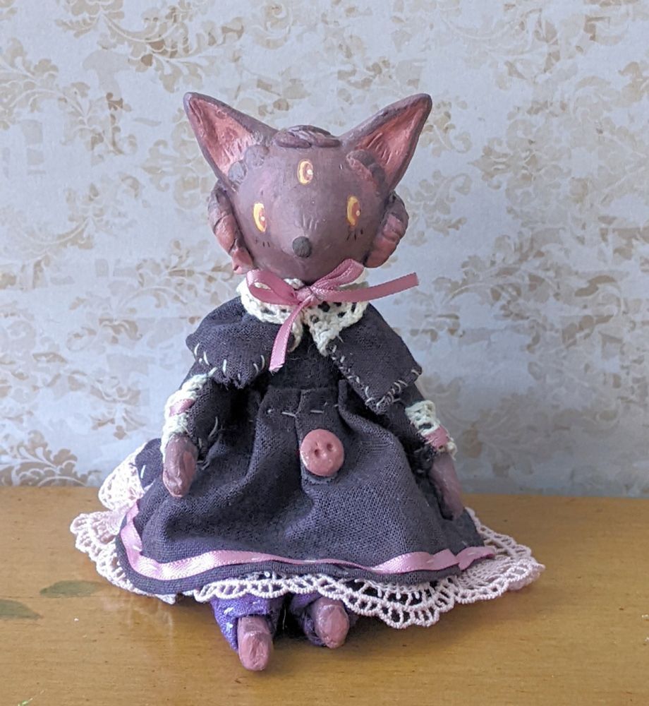 An art doll of a purple wolf character with three eyes, and horns. She's wearing a dark purpley-black dress with a cape, pink bow and lacy trim with a single pink button on front.