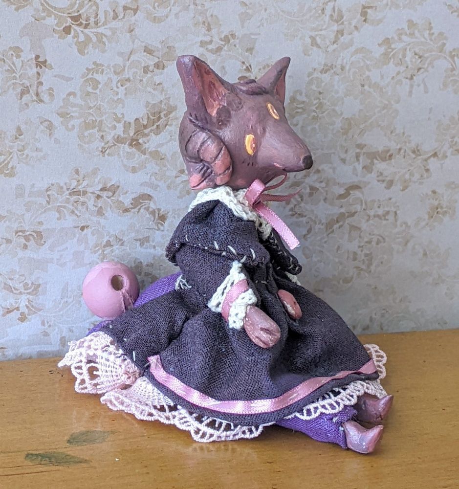 An art doll of a purple wolf character with three eyes, and horns. She's wearing a dark purpley-black dress with a cape, pink bow and lacy trim with a single pink button on front.