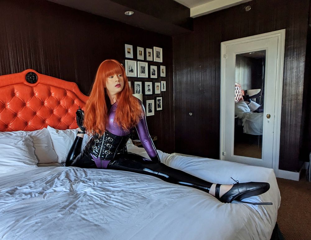 Redhead dominatrix on a bed doing a split in ballet heels and a purple and black latex outfit.