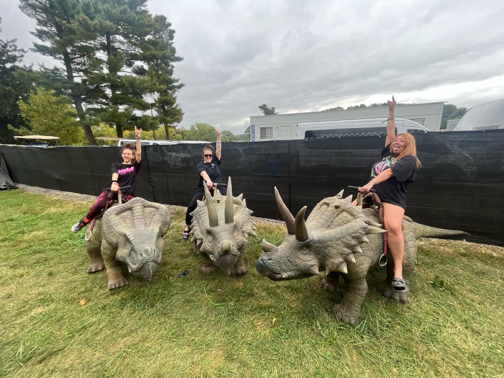 Three people are riding realistic looking triceratops “baby” dinosaurs. All three people have “rock on” hand gestures raised high into the air. 