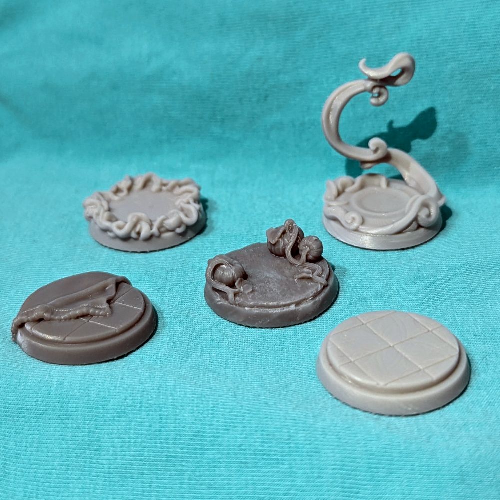 On a teal background, gray prints of bases for ttrpg miniatures. Left to right: a base depicting a tiled floor with a corner of a carpet on it, one with a twirling spell encircling it, one with pumpkins growing on it, one with a spell coiling in the air, and one depicting a tiled floor.