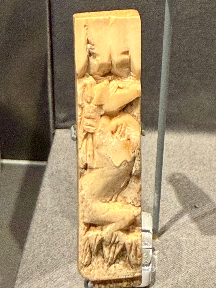 A small rectangular ivory fragment with an image of a dog headed figure holding a spear. Stylised lotus flowers can be seen towards the bottom of the fragment.