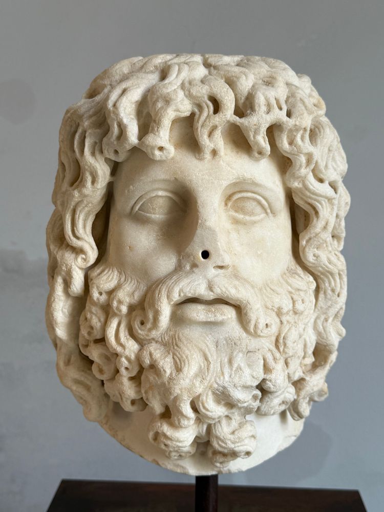 White marble statue bust of a man with long gnarly hair and shaggy beard. His nose has been broken off. 