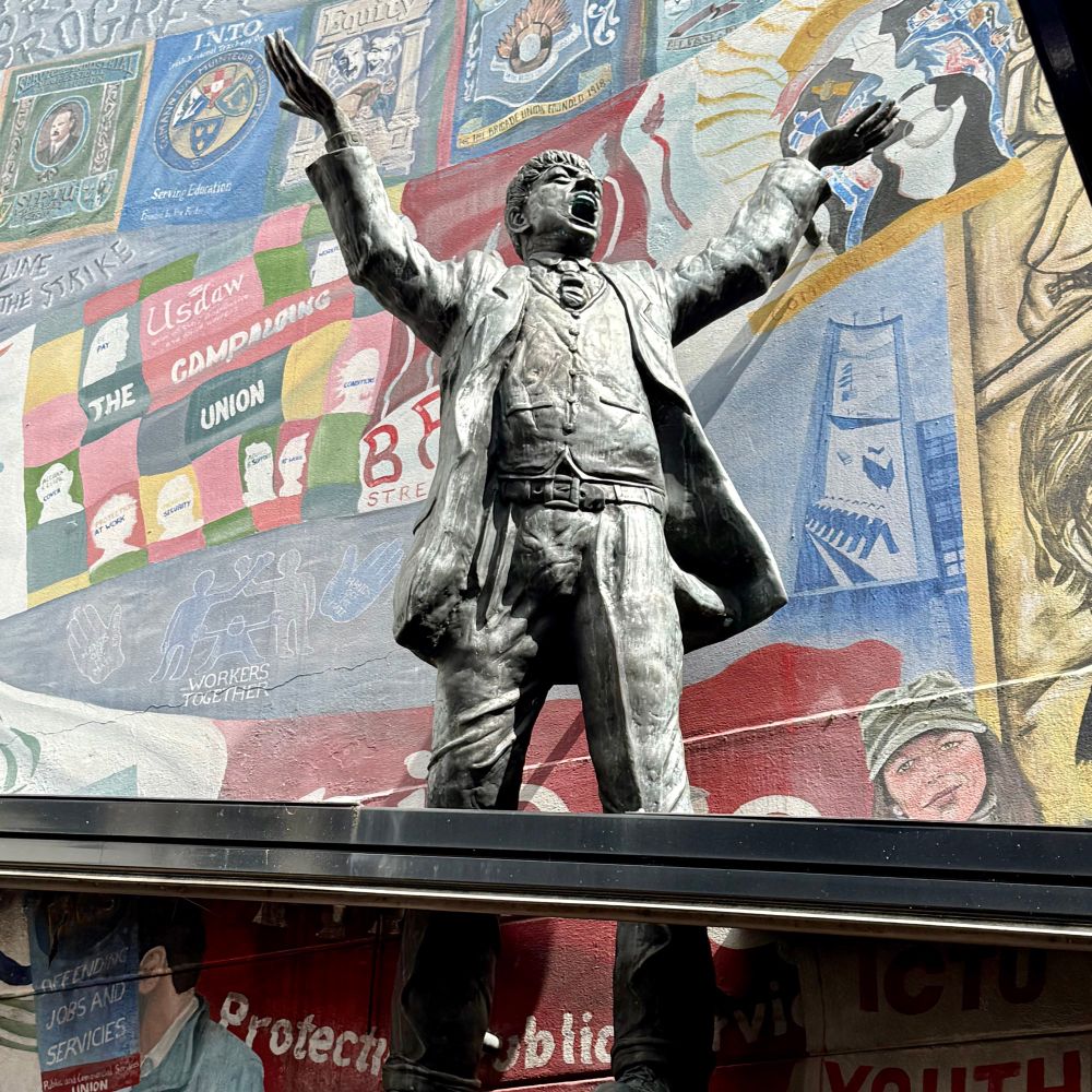 A statue of a man, whose arms are raised in exhortation and mouth is open as in speech. The statue is placed before a mural showing various union banners. 