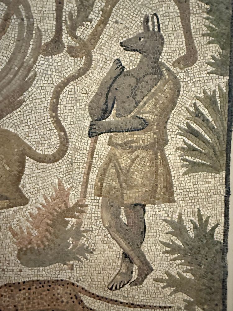 A figure with a dogs head with long snout and erect ears, recalling Anubis, wears a robe that shows off his pecks to best effect. He is stood in a landscape full of plants. Two other animals are partly visible in this photo: to his left the hind parts of a griffin and above him the legs of a giraffe.