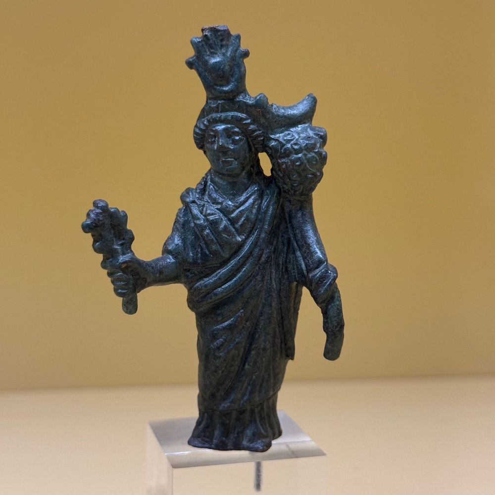 A small bronze statuette of a woman with an Egyptian solar horn headdress. She is holding a large cornucopia in her left hand and a smaller object in her right, perhaps a sistrum, torch or bouquet of roses. 
