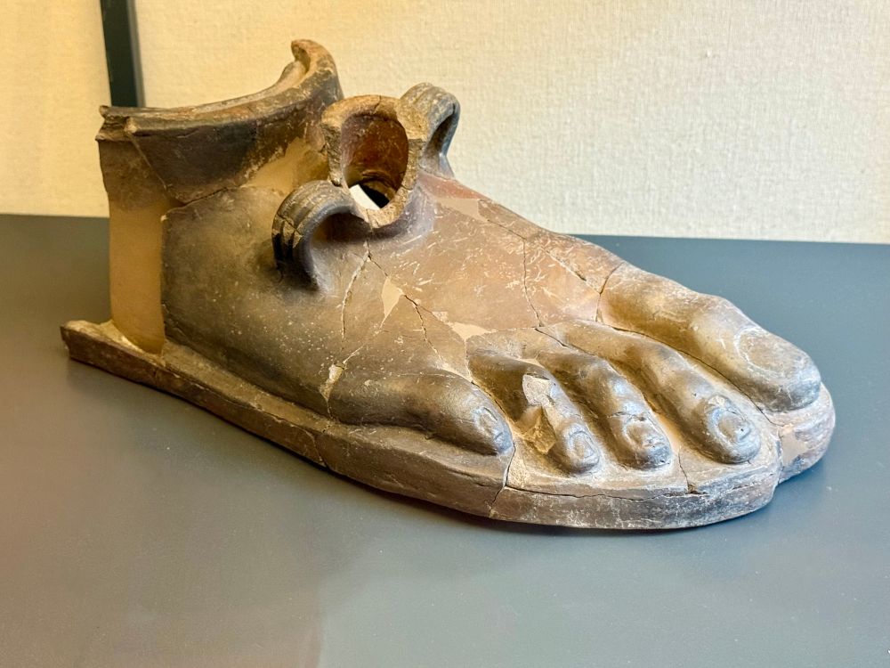 Vase in the shape of an unnecessarily detailed foot. There is a bottle mouth and handles at the top of the foot. 