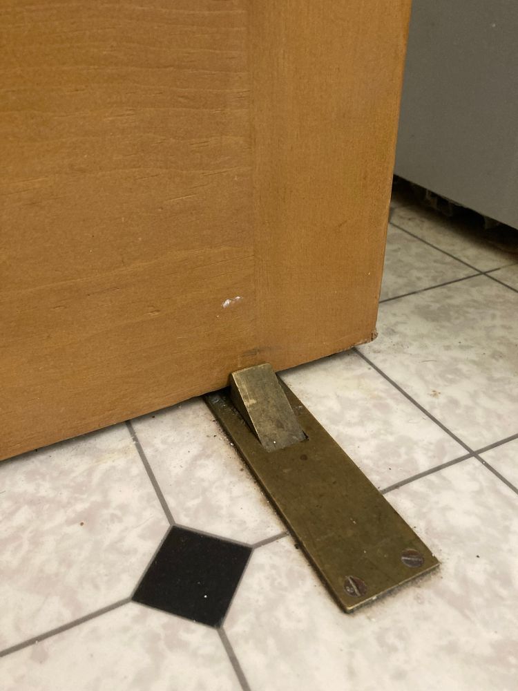 A brass doorstop holding a wooden door on a vinyl floor. 