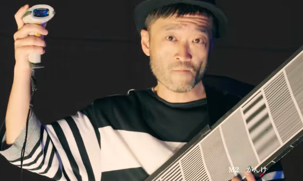 Picture of a Japanese man seen from the chest up facing the camera, staring right at it with a calm serious expression. The background is black. He is wearing a shirt covered in black and white stripes of varying widths, and his left hand is cradling a long rectangular board with black-on-white striped markings. His right arm is raised, brandishing a wired barcode reader.