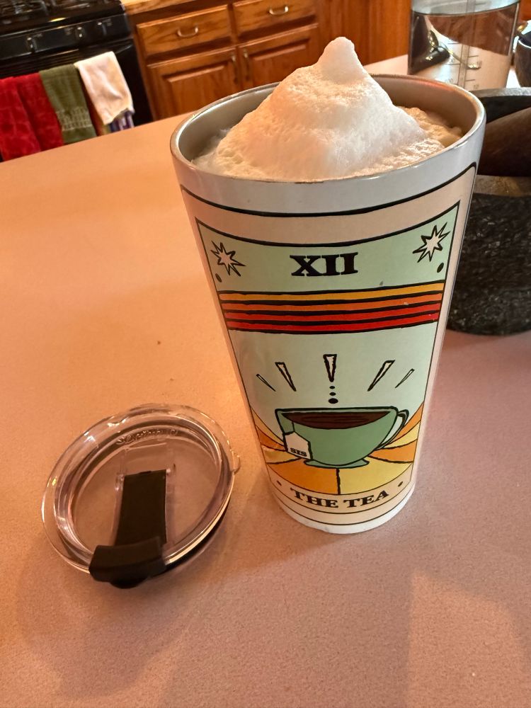 A thermal cup with “The Tea” tarot card design, containing a foaming latte drink. 