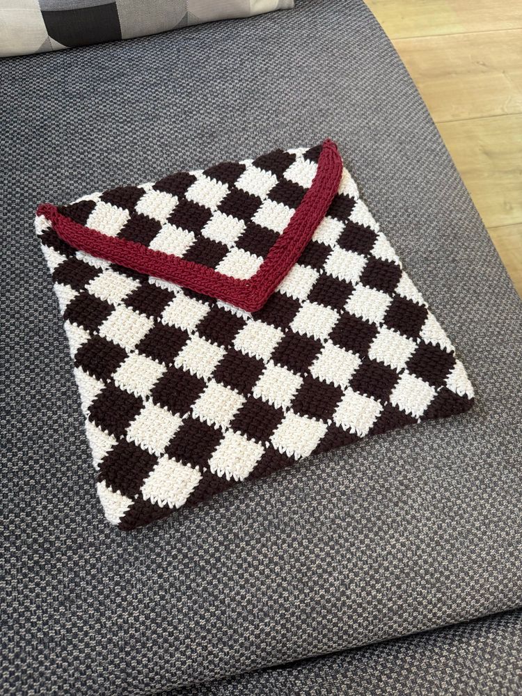 Handmade crochet envelope bag with a checkered pattern of black and white squares, finished with a red triangular flap. The bag is displayed on a grey textured cushion.