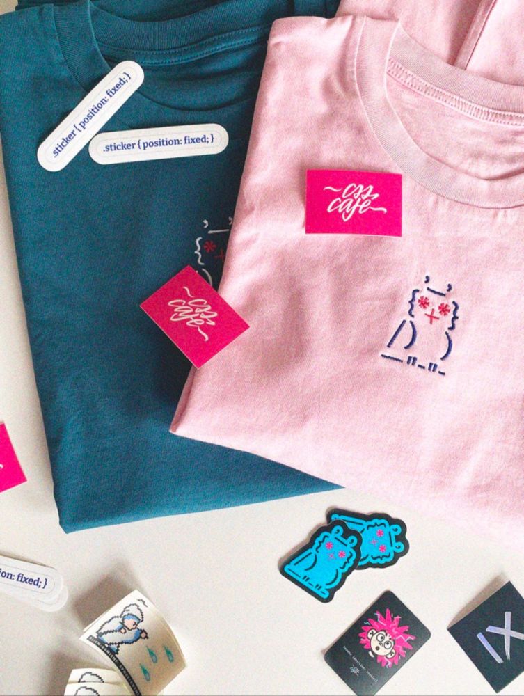 Two folded T-shirts lie side by side on a white surface — one teal, one light pink. Both shirts feature an embroidered version of the "lobotomized owl," a quirky character by accessibility advocate Heydon Pickering, with red star-shaped eyes and a stitched mouth. Scattered around are various colorful stickers: two bright pink ones with the text “CSS Café,” two white ones styled like CSS code that read “.sticker { position: fixed; }”, and several playful character stickers, including blue creatures, raindrops, and a spiky-haired pink face. There’s also a small roll of stickers in the corner. The scene conveys a fun, creative atmosphere with a strong web developer and accessibility culture vibe.