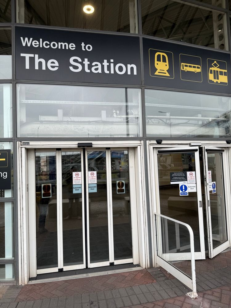 Entrance to a modern transportation hub labeled “Welcome to The Station,” featuring icons for train, bus, and tram services above glass automatic doors.