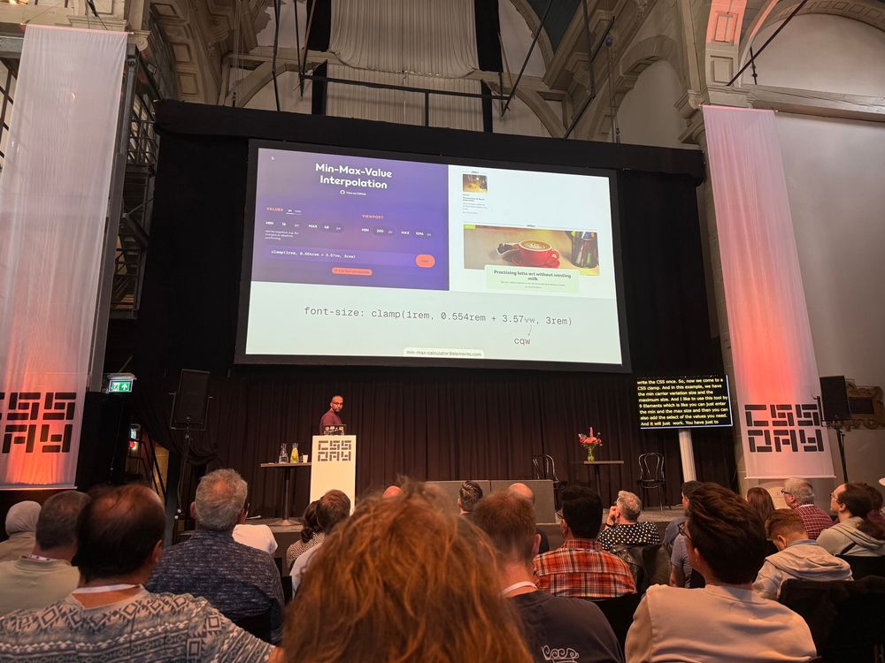 Ahmad Shadeed speaking on stage at CSS Day in a large hall with high ceilings and classical architecture. A large slide behind him explains min-max-value interpolation in CSS using the clamp() function. The audience is seated and attentively watching the talk. Captions are displayed on a screen in the lower-right corner of the stage.