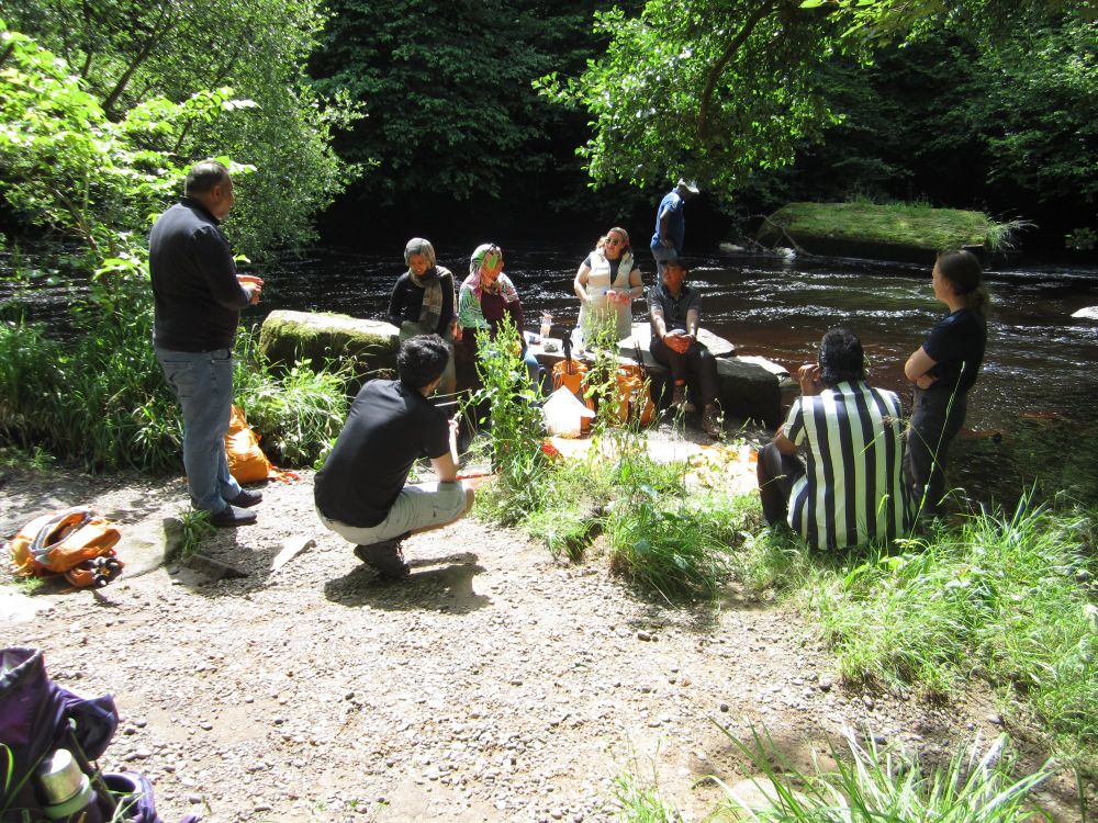 A group of hikers have lunch on a riverbank next to a woodland trail.