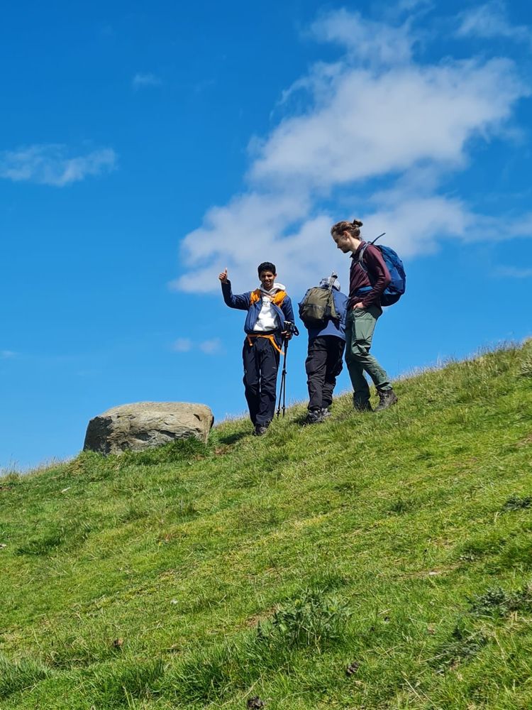 Three hikers on a steep grassy slope next to a large boulder.