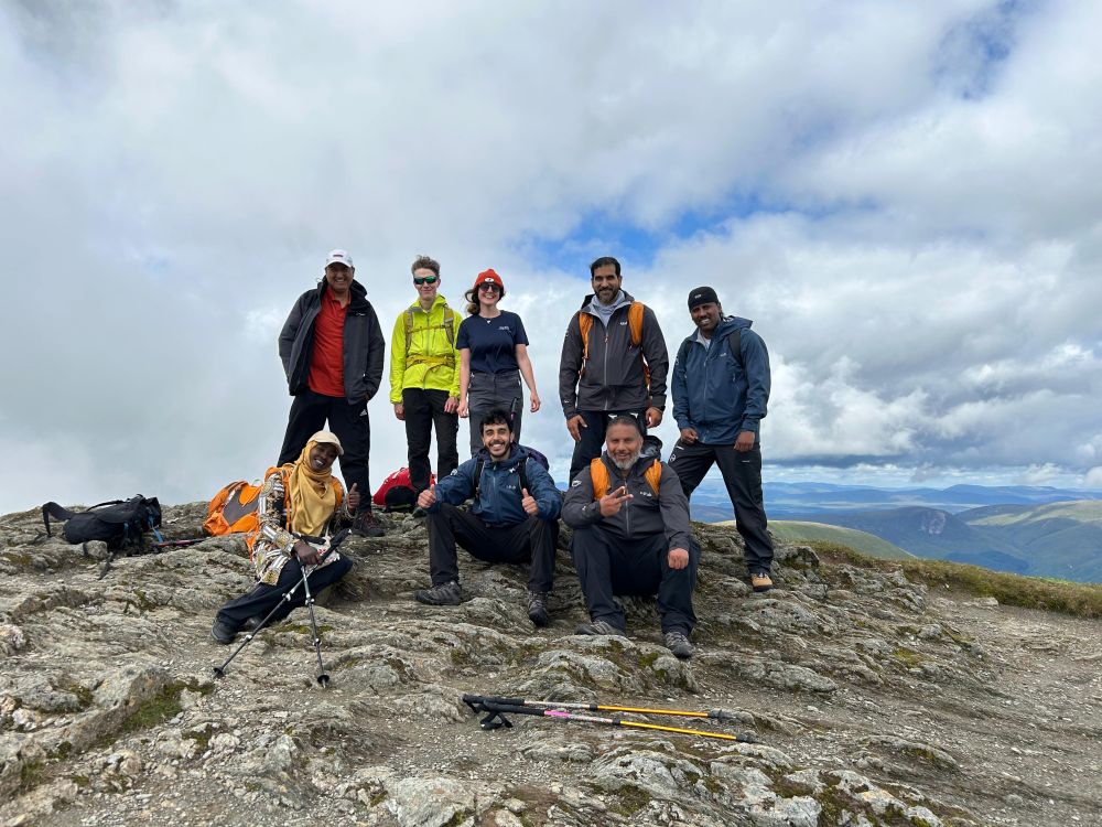 A group of eight hikers at the summit of a mountain with large clouds overhead and mountains in the distance.
