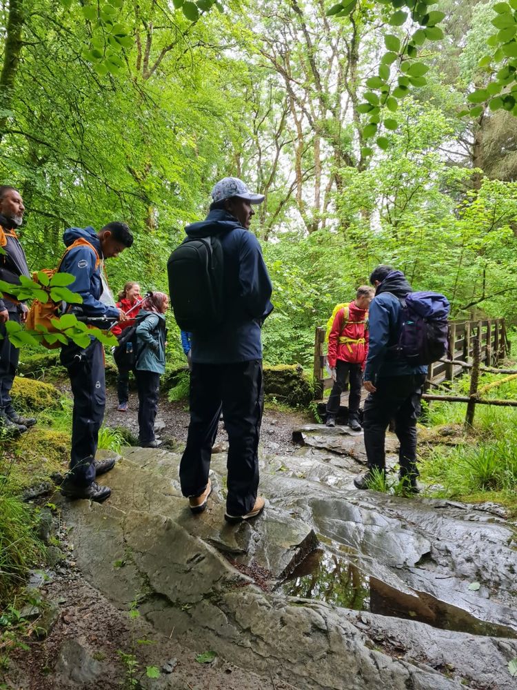 A group of hikers standing on a wet, rocky forest trail. There are trees all around and a wooden footbridge in the background.
