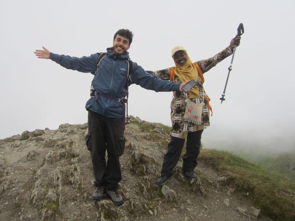 Two hikers celebrate with arms out wide and big smiles at the summit of a mountain.