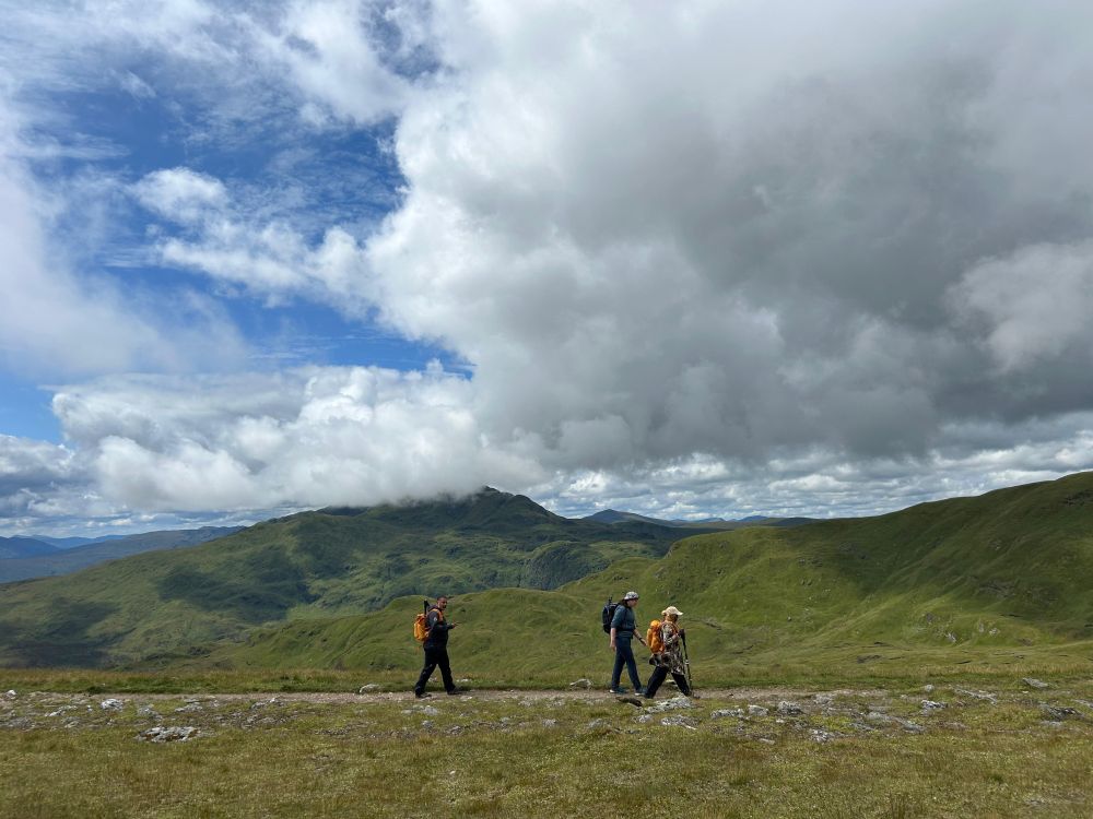 Three hikers stride across a flat path with mountains in the background and large clouds gathering above.