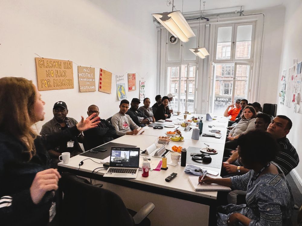 A woman delivers a presentation to a group seated around a long table, in a white room with large windows at the back. On the walls are various signs, including ‘GLASGOW HAS NO PLACE FOR FASCISTS’ and ‘NO HOSTILE ENVIRONMENT IN SCOTLAND’.