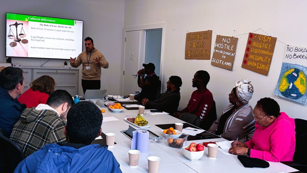 A man standing in front of a large screen delivers a presentation for a group seated around a long table. On the walls are various signs, including 'GLASGOW HAS NO PLACE FOR FASCISTS' and 'NO HOSTILE ENVIRONMENT IN SCOTLAND'. The screen shows a slide headed 'Cultural Differences', with the subheading 'The Rule of Law: Justice for All'.