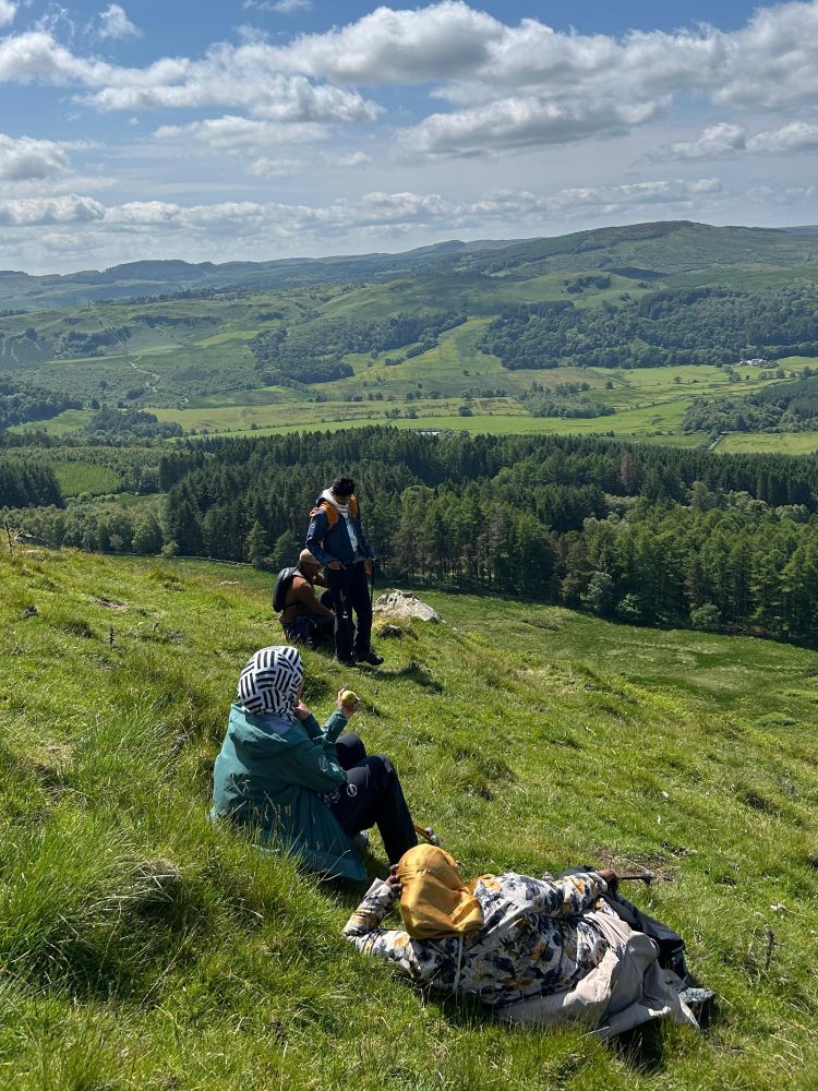 A group of hikers sitting on a grassy slope, enjoying the view of hills and trees in the distance.