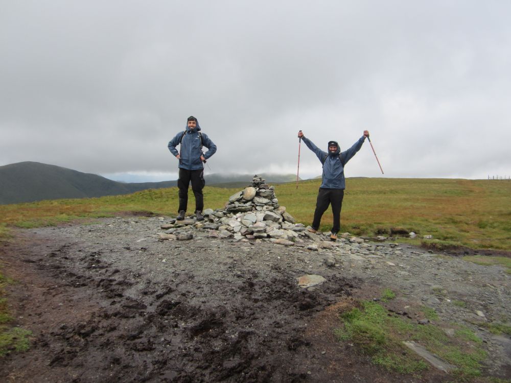 Two hikers standing either side of a cairn at the summit of a hill. Behind them is another mountain with low cloud moving in.