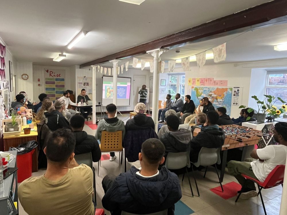 A seated audience of 25 in a large, brightly-lit room, watching a presentation delivered by a member of the Community InfoSource outreach team.