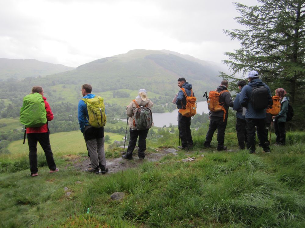 A group of hikers stand at a viewpoint with backs to the camera, looking across to a loch and distant hills.