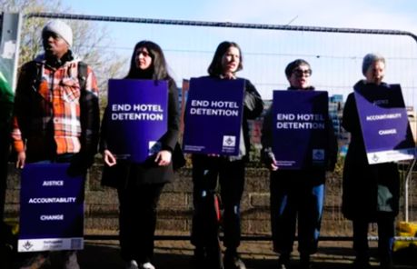 Five protesters standing together holding placards reading, "JUSTICE ACCOUNTABILITY CHANGE", and "END HOTEL DETENTION".