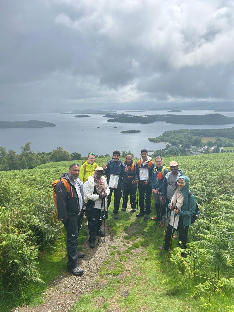 A group of nine hikers pose for a photo standing on a path surrounded by fern. In the background is a loch with several small islands and thick clouds overhead.