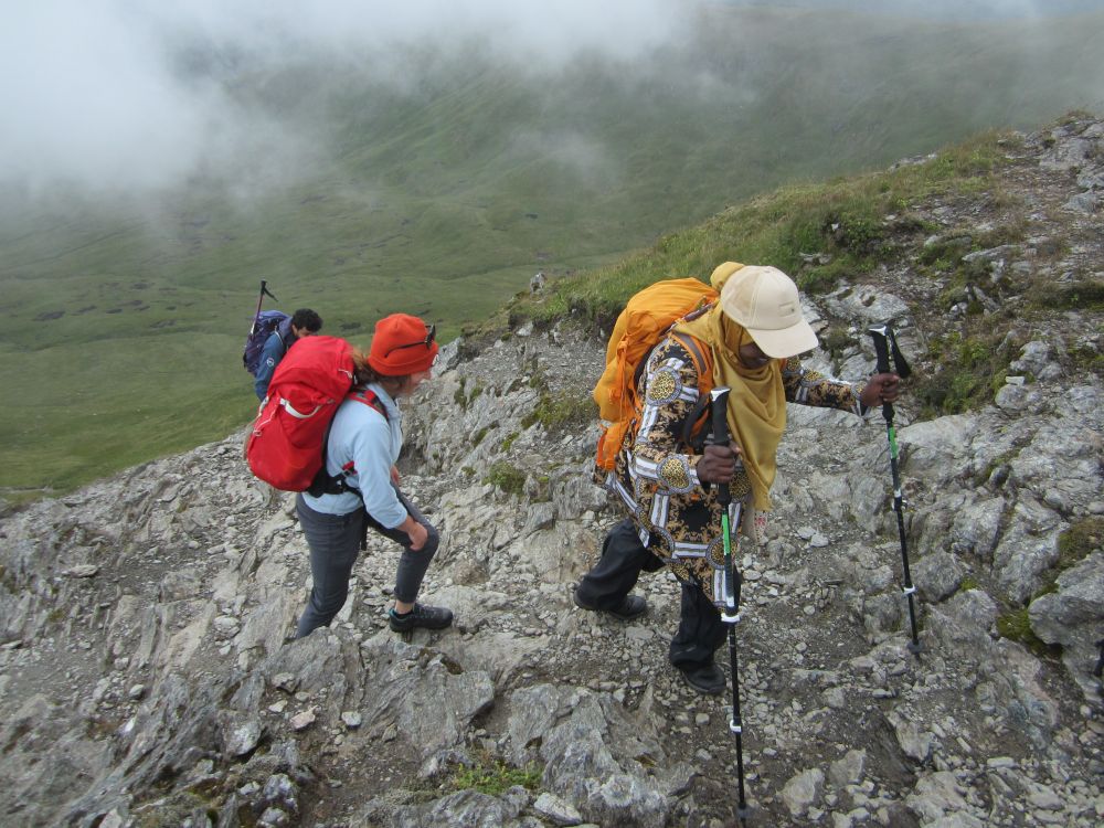 Three hikers climb a rocky path as low cloud drifts in.