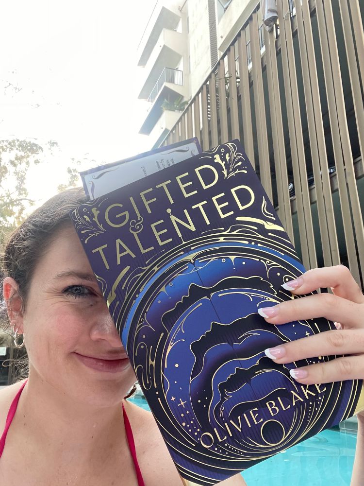 A selfie of me in a pool holding up the Fairyloot edition of Gifted and Talented by Olivie Blake, which has a cover with gold foiled circular designs with purple and blue 